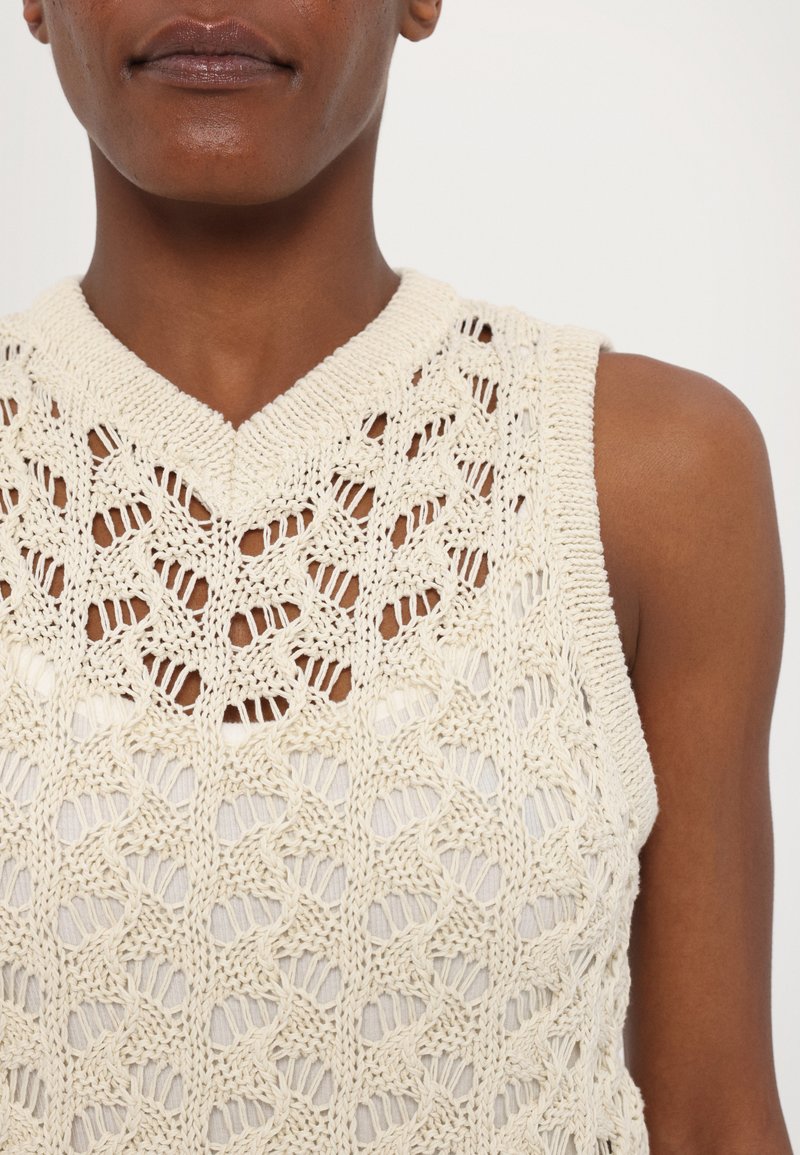 Débardeur tricoté sans manches en fil couleur crème avec un motif complexe ; présente un col en V et un design en maille ouverte.