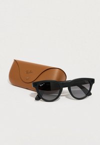 Gafas de sol Ray-Ban negras con forma redonda y lentes oscuras, acompañadas de un estuche de cuero marrón. Presentan un pequeño detalle de logo en la montura.