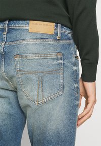 Jeans in denim blu con una finitura leggermente consumata. Presentano un'etichetta in pelle marrone con la scritta "TIGER OF SWEDEN" e cuciture arancioni sui taschini.