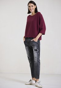 Burgunderfarbener Strickpullover mit lockerer Passform und verkürztem Design, kombiniert mit zerrissenen grauen Jeans und weißen Sneakers mit schwarzen Akzenten.
