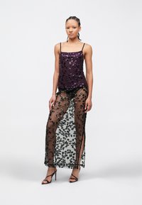 Vrouw met gevlochten haar, gekleed in een paarse top met pailletten en een lange, doorschijnende zwarte rok met bloemenpatronen, gecombineerd met zwarte sandalen met hoge hakken.