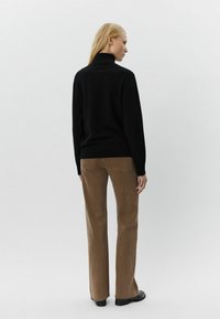 Maglione nero a collo alto con orlo a costine abbinato a pantaloni marroni a gamba dritta. Il modello è in posizione di spalle rispetto alla fotocamera, con capelli lisci e biondi.