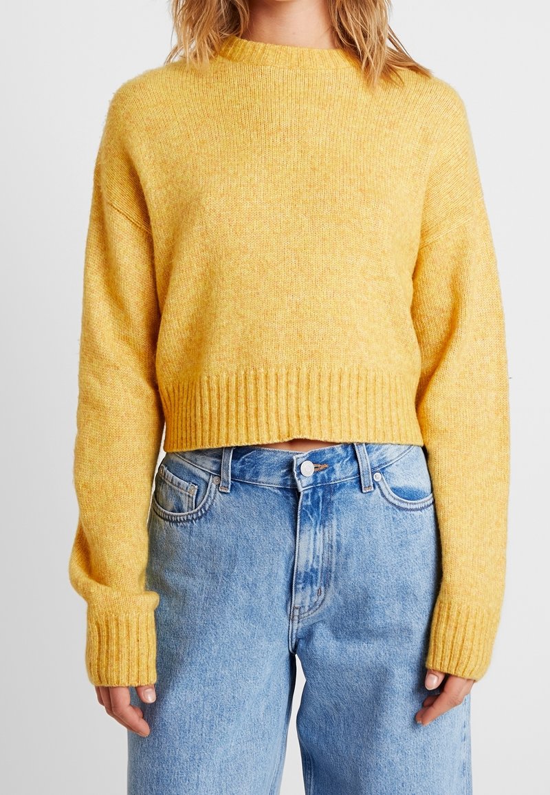 Femme portant un pull jaune à manches longues col rond et un jean bleu clair taille haute, debout avec les bras relâchés le long du corps.