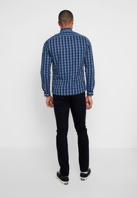 Chemise boutonnée à motif à carreaux bleus, avec manches longues et col. Vue de dos montrant une coupe ajustée et un pantalon noir contrastant.