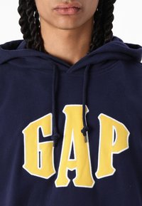 Hanorac bleu marin, cu un logo mare galben "GAP", cu o textură moale, sireturi și un design clasic de tip pulover.