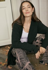 Zwarte gebreide cardigan met een losse pasvorm, gedragen over een wit topje. Het model zit op een stoel en draagt een broek met een patroon met vloeiende roze ontwerpen.