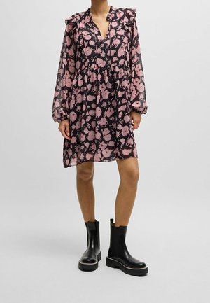 Frau in einem schwarzen und rosa geblümten Kleid mit langen Ärmeln, die mit den Händen seitlich steht, kombiniert mit schwarzen Lederknöchelstiefeln.