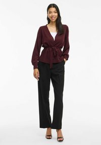 Cardigan a maglia Bordeaux con scollo a V, cintura da annodare in vita e maniche lunghe, abbinato a pantaloni neri a gamba larga e décolleté neri con punta aperta.