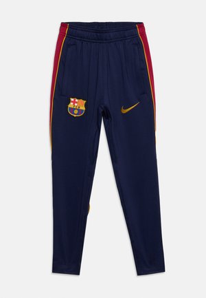 Granatowe sportowe spodnie FC Barcelona z czerwono-żółtymi paskami po bokach, elastyczną talią, herbem klubu na lewej udzie oraz złotym logo Nike na prawym udzie.