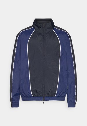 Námornícky vetrovka s bielym lemovaním, vysokým golierom, predným zipsom a elastickými manžetami. Je vyrobená z hladkej, ľahkej tkaniny.