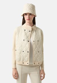 Kremowa pikowana kurtka z prążkowanym kołnierzem i zatrzaskami, w zestawie z pasującym kapeluszem typu bucket. Gładka textura i lekka konstrukcja.