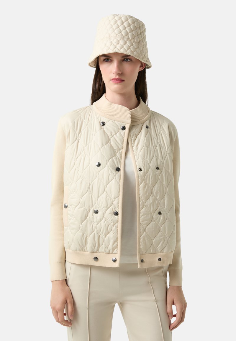 Kremowa pikowana kurtka z prążkowanym kołnierzem i zatrzaskami, w zestawie z pasującym kapeluszem typu bucket. Gładka textura i lekka konstrukcja.