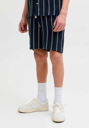 Mann trägt marineblaue Shorts und passendes Hemd mit weißen vertikalen Streifen, weiße Sportsocken und weiße Turnschuhe.