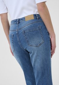 Persona vistiendo unos jeans de mezclilla azul de talle alto con bolsillos traseros y una camiseta blanca, mostrado desde la cintura hacia abajo en un ángulo.