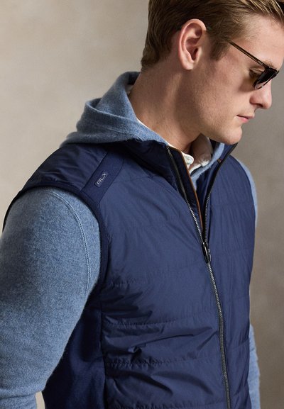 Άνδρας με γυαλιά ηλίου, μπλε μπουφάν με επένδυση πάνω από ανοιχτόχρωμο μπλε φούτερ, κοιτάζει προς το πλάι μπροστά σε απλό φόντο.