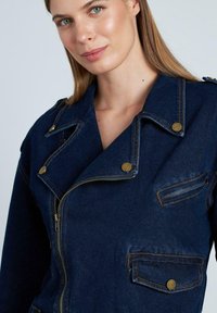 Chaqueta de mezclilla en azul oscuro con cierre de cremallera, solapas en punta y herrajes en tonalidad dorada. Presenta bolsillos en el pecho y laterales con costura superior.