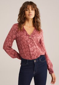 Långärmad blus med rosengrönt mönster i dämpad rosafärg, med v-ringning och knapprad vid bröstet, tillsammans med mörka denimjeans.