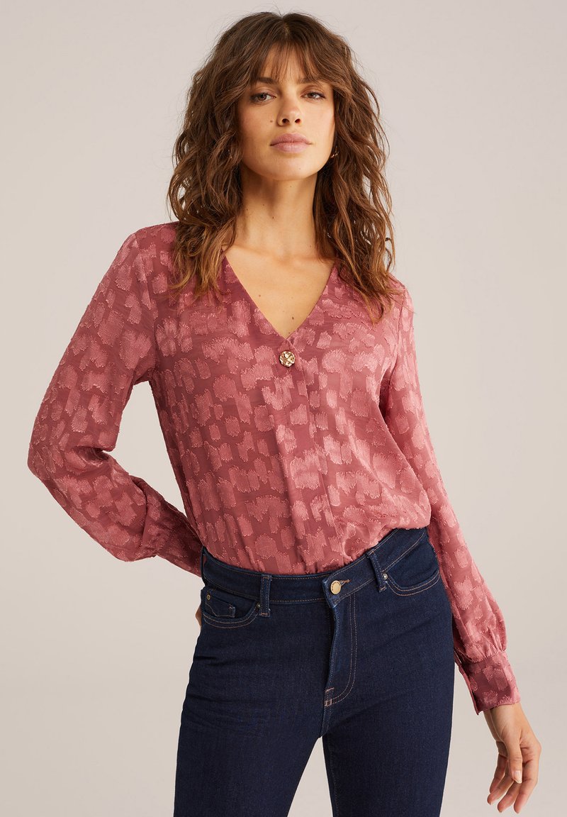 Långärmad blus med rosengrönt mönster i dämpad rosafärg, med v-ringning och knapprad vid bröstet, tillsammans med mörka denimjeans.