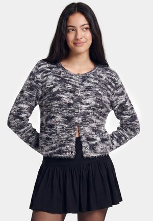 Ung kvinde med langt mørkt hår iført en grå mønstret knapfront cardigan og et sort plisseret skørt, smilende og kigger til siden.