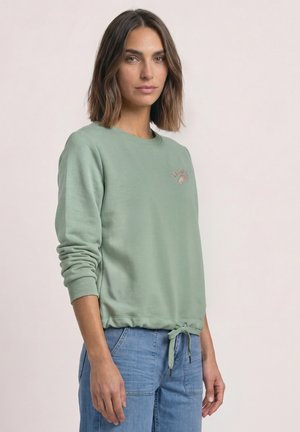 Kvinde med skulderlangt brunt hår iført salviegrøn sweatshirt med pink tekst og croissant-grafik, kombineret med lyseblå jeans.