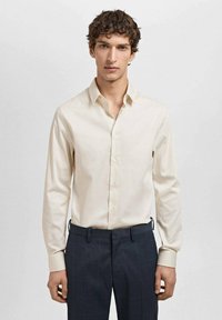 Cămașă lungă, bej deschis, cu guler cu nasturi, textură netedă și design ajustat, asociată cu pantaloni închiși, cu carouri.