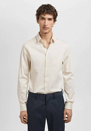 Ljust beige långärmad skjorta med knapp-down krage, slät textur och åtsittande design, tillsammans med mörka, rutiga byxor.