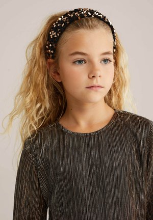 Bandă de cap neagră cu paiete și accente strălucitoare, asociată cu un top negru texturat cu dungi metalice verticale. Păr lung, ondulat, blond.