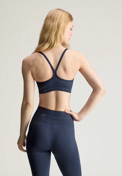 Woman with long blonde hair wearing a dark blue sports bra and matching leggings, facing away with one hand on her hip.