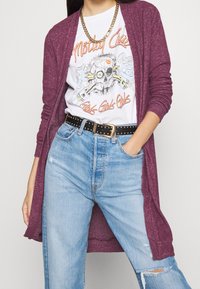 Plommonfärgad lång cardigan över en vit t-shirt med grafiskt tryck som har ett dödskallemotiv, matchad med ljusblå slitna jeans och ett nitarbälte i svart.