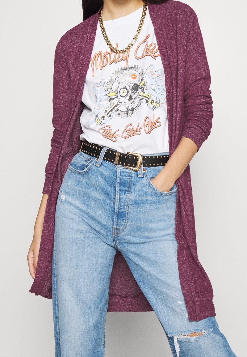 Plommonfärgad lång cardigan över en vit t-shirt med grafiskt tryck som har ett dödskallemotiv, matchad med ljusblå slitna jeans och ett nitarbälte i svart.