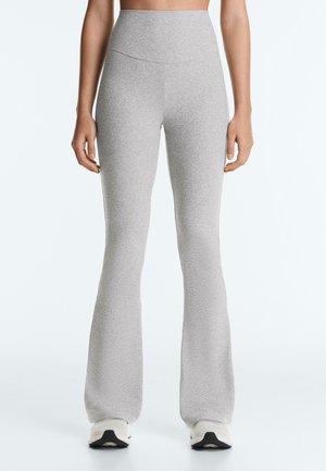 Femme portant un pantalon évasé gris clair taille haute et des baskets blanches, debout devant un fond blanc uni.