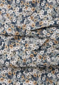 Close-up van een opgevouwen shirt met een klein, multikleurig bloemenpatroon in gedempt blauw, grijs, wit en tan, met zichtbare knopen en knoopsgaten.
