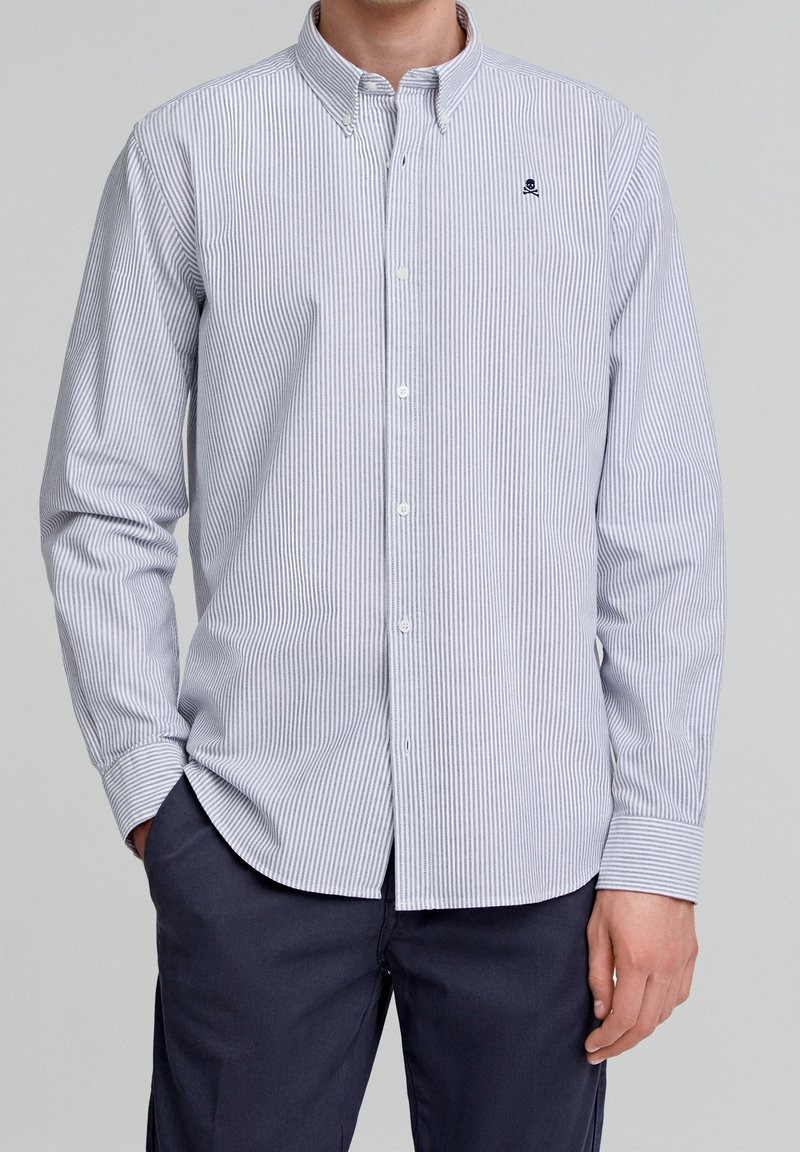Homme portant une chemise à rayures verticales bleu clair et blanc avec un petit logo de crâne brodé sur la poitrine, associée à un pantalon bleu foncé.