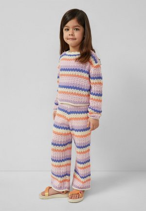 Junges Mädchen steht, trägt einen pastellgestreiften Strickpullover und passende Hosen mit orange-beigen Sandalen, vor einem schlichten grauen Hintergrund.