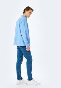 Camisa a rayas de color azul claro con cierre de botón, combinada con jeans azul oscuro y zapatillas blancas. La camisa tiene un ajuste relajado y mangas largas.