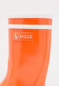 Pomarańczowy kauczukowy kalosz z białymi akcentami i etykietą logo. Charakteryzuje się gładką, błyszczącą powierzchnią oraz zaokrąglonym czubkiem.