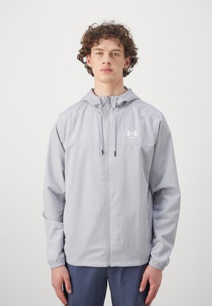Casaco corta-vento cinza claro com capuz, fecho de correr frontal, punhos elásticos e logótipo Under Armour no peito. Tecido liso e leve.