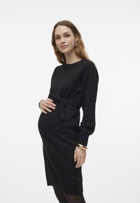 Black knitted dress with long sleeves, cinched waist, side tie, and ribbed hem. Model poses, highlighting belly and simple gold bracelet.