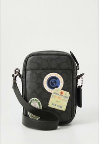 Bolso bandolera de cuero negro con un patrón texturizado, con pegatinas de temática de viaje y una correa desmontable. Forma rectangular con cremalleras.