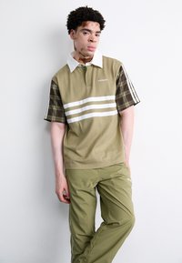 Grün- und braunkariertes Kurzarm-Polohemd mit weißen Streifen, kombiniert mit passenden olivgrünen Hosen. Adidas-Logo auf der Brust und an den Hosen.