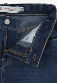 Jeans ajustados de denim azul oscuro con cremallera de latón, cierre de botón y costuras amarillas en contraste. La etiqueta de tamaño dice "12."