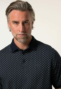 Laivastonsininen polo-paitoja, jossa on vaaleita geometrisia kuvioita. Varustettu kiinteällä kauluksessa, kolmella napilla varustetulla avauskannella ja pienellä logolla rinnassa.