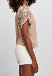 Braunes kurzärmliges Oberteil mit Spitzenakzenten an den Schultern, glatter Textur und lockerer Passform. Kombiniert mit weißen Shorts mit umgeschlagenem Saum.