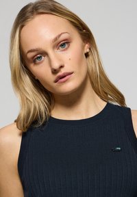 Tajt, ärmlös topp i mörk marinblå ribbad tyg med rund halsringning och en liten grön detalj på vänster bröst.