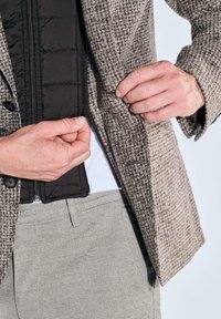 Braunes Hahnentritt-Sakko mit schwarzer, gesteppten Innenfütterung. Verfügt über zwei Knöpfe, verstellbaren Reißverschluss und sichtbare Nähte. Hellgraue Hose.