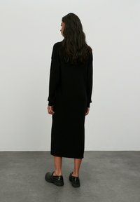 Schwarzes, geripptes Strickkleid mit langen Ärmeln und knielanger Länge, kombiniert mit klobigen schwarzen Schuhen. Glatte Textur, schlichtes Design.
