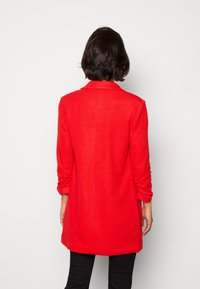 Blazer largo rojo de tela texturizada. Presenta un cuello con muescas y mangas enrolladas. La parte posterior es ligeramente más larga con un corte recto.