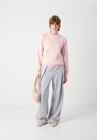Ljusrosa bomulls-sweatshirt med rund halsringning och grön logotyp, kombinerad med grå vida sweatpants och en beige axelväska.