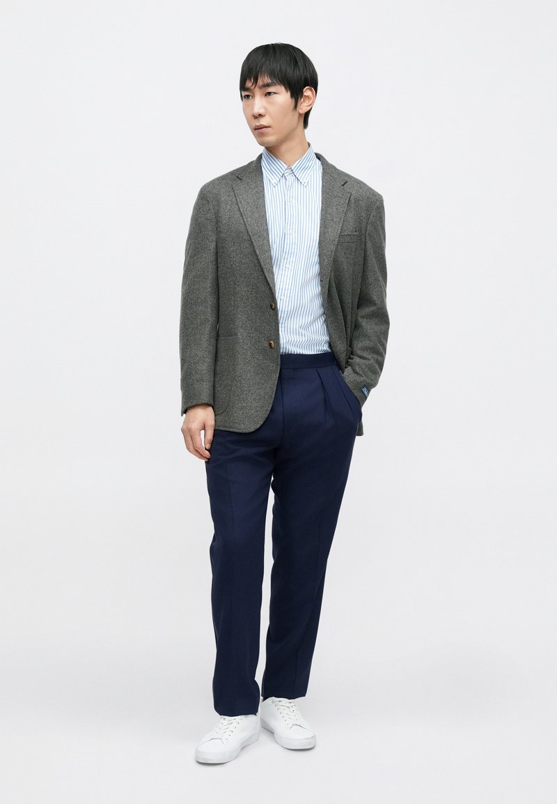 Grijze wollen blazer over een lichtblauw gestreept overhemd, marineblauwe broek en witte sneakers. De blazer heeft een borstzakje en knopen.