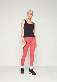 Naine musta sporditopiga, koralli värvi legginsite ja mustade tossudega, hoiab valget veepudelit käes, seistes ühetoonilise heleda tausta ees.